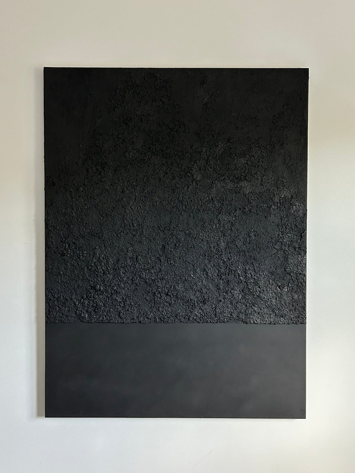 A minimalist black textured wall art panel mounted on a light gray wall. The panel features a two-tone design with a heavily textured dark upper half and a smooth matte black lower half.
