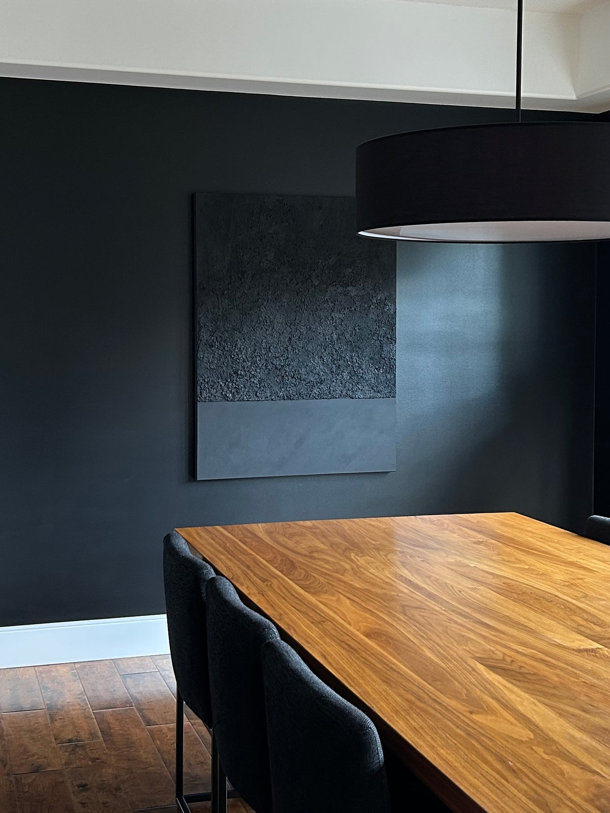 A modern dining room setup featuring a rectangular wooden dining table with black chairs, a black drum pendant light, and a prominent dark charcoal wall in a home interior.